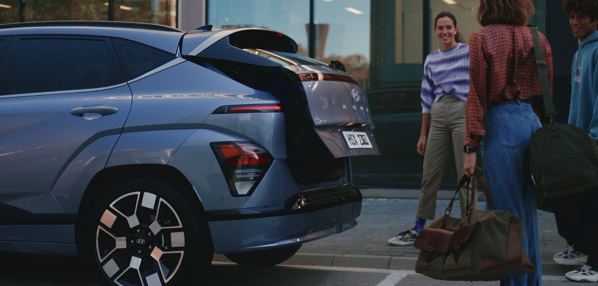 A green The all-new KONA is parked on the side of the road. A woman holding a box in both hands is standing behind The all-new KONA, and the trunk of The all-new KONA is being opened.